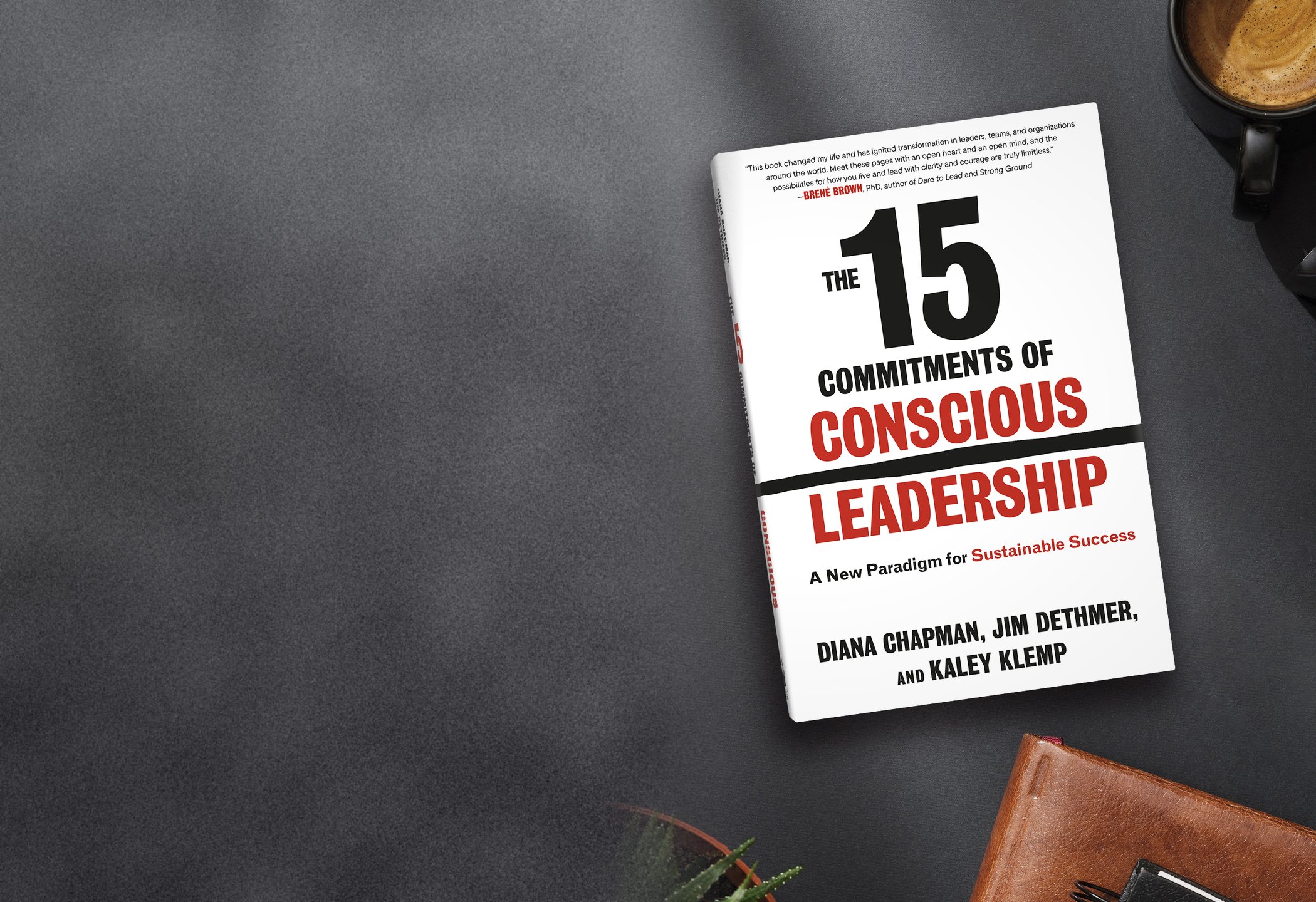 The 15 Commitments of Conscious Leadership hardcover lying on a slate desk beside an espresso, a leather journal, and a small succulent.